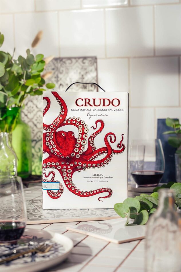 Crudo Nero D'Avola Cabernet Sauvignon bag-in-box and wine glass arranged on a dining table. Product image.